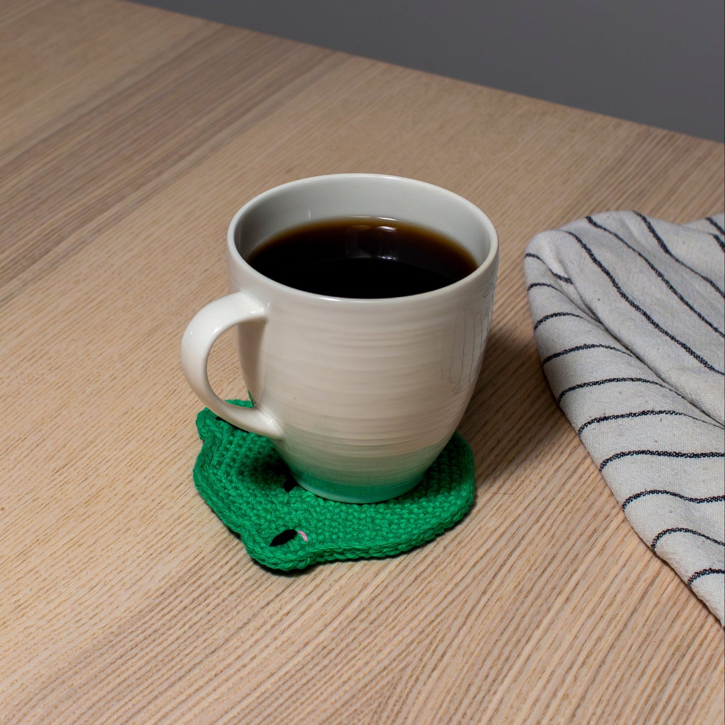 Vihreä käsintehty virkattu sammakkoa esittävä lasinalunen tai mukinalunen vaalealla pöydällä, jonka päällä valkoinen muki, jossa kahvia sekä vieressä keittiöliina.