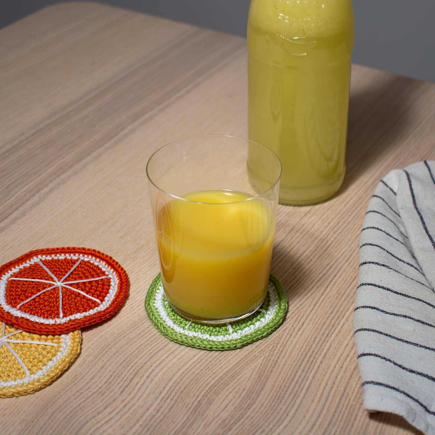 Vihreä käsintehty virkattu lime lasinalunen tai mukinalunen pöydällä, jonka päällä lasi jossa appelsiinimehua keittiöliinan ja keltaisen sitruuna sekä oranssin appelsiini lasinalusten kanssa.
