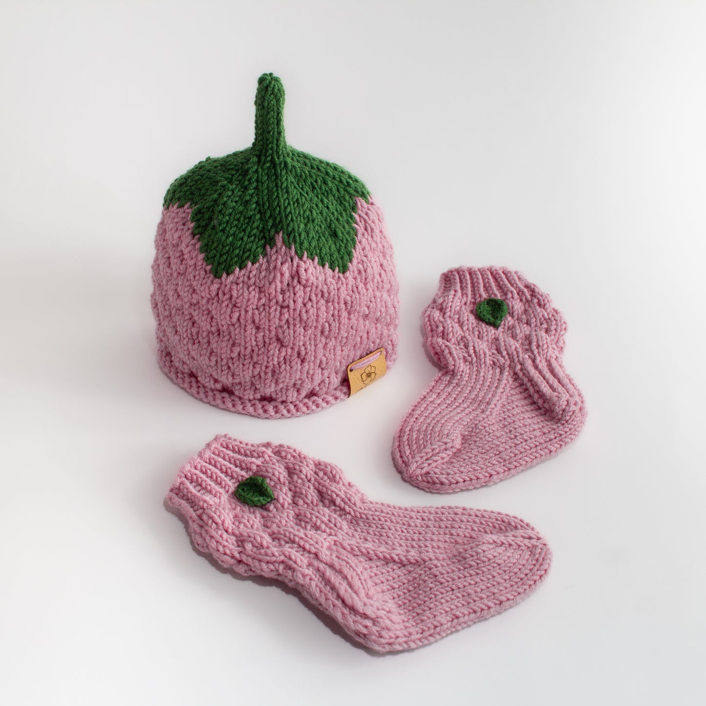 Vaaleanpunainen käsintehty neulottu vadelmapipo, jossa vihreä lehtiosuus sekä kaksi pientä vauvan vaaleanpunaista vadelma villasukkaa, jossa pienet lehdet.