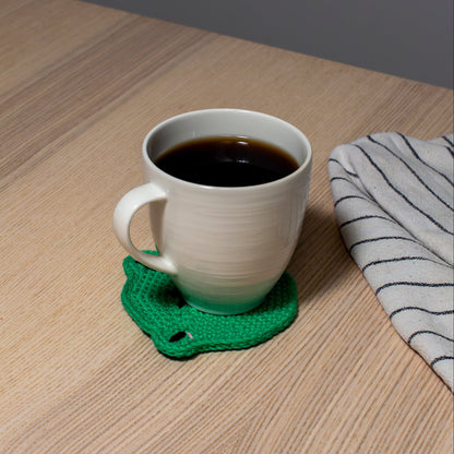 Vihreä käsintehty virkattu sammakkoa esittävä lasinalunen tai mukinalunen vaalealla pöydällä, jonka päällä valkoinen muki, jossa kahvia sekä vieressä keittiöliina.
