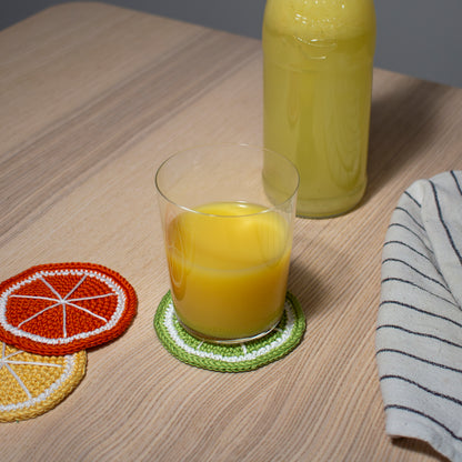 Vihreä käsintehty virkattu lime lasinalunen tai mukinalunen pöydällä, jonka päällä lasi jossa appelsiinimehua keittiöliinan ja  keltaisen sitruuna sekä oranssin appelsiini lasinalusten kanssa.