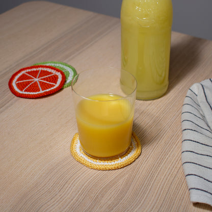 Keltainen virkattu sitruuna lasinalunen tai mukinalunen pöydällä, jonka päällä lasi jossa appelsiinimehua keittiöliinan ja appelsiini sekä lime lasinalusten kanssa.