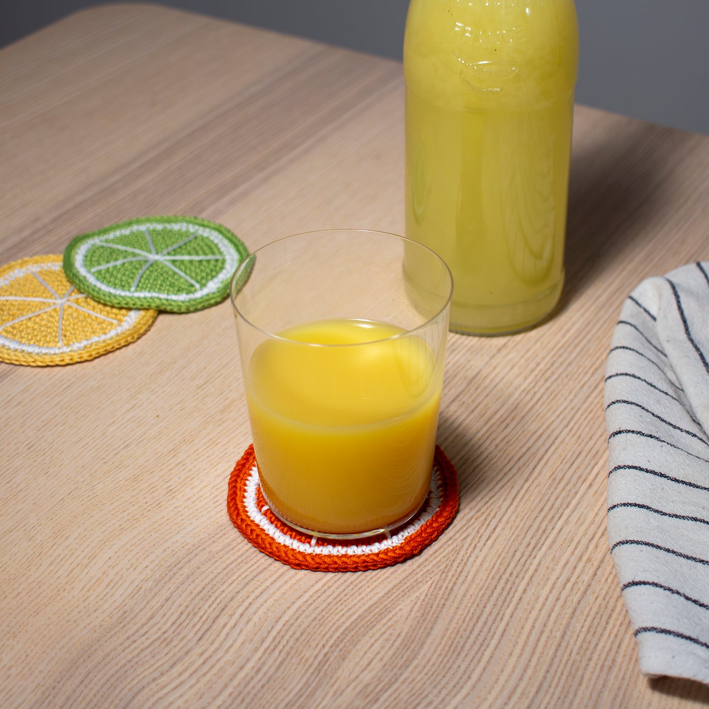 Oranssi virkattu appelsiini lasinalunen tai mukinalunen pöydällä, jonka päällä lasi jossa appelsiinimehua keittiöliinan ja  keltaisen sitruuna sekä vihreän lime lasinalusten kanssa.